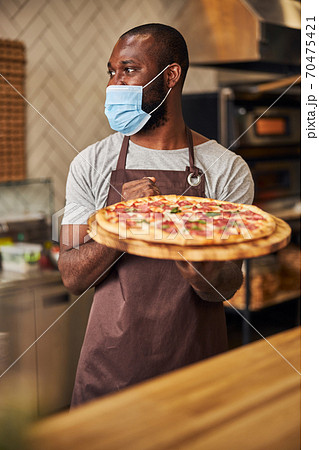 Afro American man in protective face mask holding pepperoni pizza Afro American man in protective face mask holding pepperoni pizza 70475421