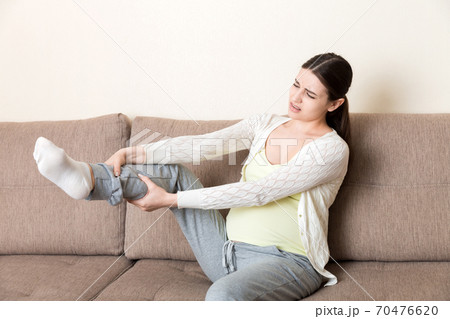Leg cramps during pregnancy. Closeup of hands massaging swollen foot while sitting on sofa Leg cramps during pregnancy. Closeup of hands massaging swollen foot while sitting on sofa 70476620