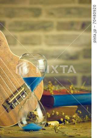 A vintage hourglass and ukulele with an old book and brass pen on a wooden table and brick background in the morning. Closeup and copy space. The concept of memories or things in the past. 70480920
