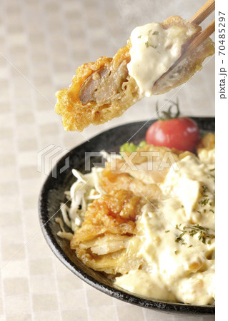 ミニトマトなど野菜も一緒に乗っているタルタルソースたっぷりのチキン南蛮丼の一切れを箸でつまんで食べる 70485297