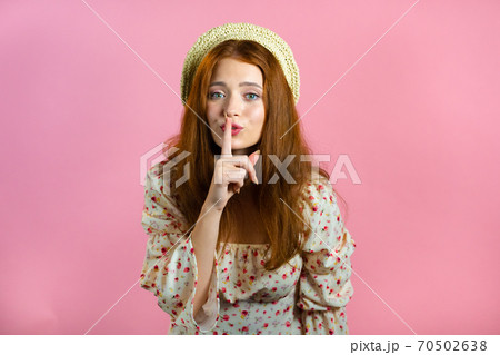 Smiling woman with red hair holding finger on her lips over pink background. Gesture of shhh, secret Smiling woman with red hair holding finger on her lips over pink background. Gesture of shhh, secret 70502638