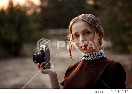 close-up portrait of a young beautiful girl in a brown dress in a retro style with a vintage camera in her hands on an abandoned road 70516514