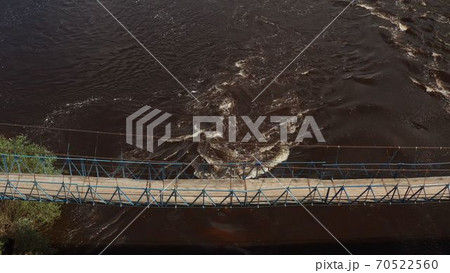 Suspended footbridge across the river. Suspended wooden pedestrian bridge across the river. bridge in the forest. green trees, foliage of trees, summer days.old hanging footbridge across a small river 70522560