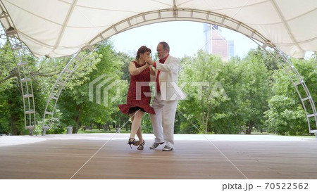 Accordion and violin playing in the park. A woman in a red dress and a man in a white suit are dancing tango. An elderly couple dancing tango in the park. 70522562