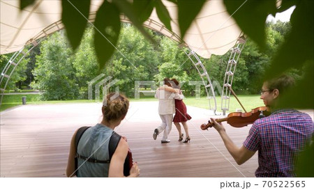 Accordion and violin playing in the park. A woman in a red dress and a man in a white suit are dancing tango. An elderly couple dancing tango in the park. Accordion and violin playing in the park. A woman in a red dress and a man in a white suit are dancing tango. An elderly couple dancing tango in the park. 70522565