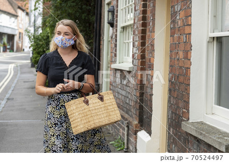 Young blond woman wearing face mask walking through village, carrying shopping bag. Young blond woman wearing face mask walking through village, carrying shopping bag. 70524497