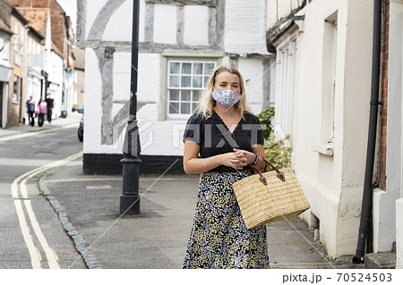 Young blond woman wearing face mask walking through village, carrying shopping bag. Young blond woman wearing face mask walking through village, carrying shopping bag. 70524503