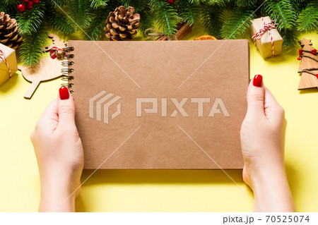 Top view of female hand making some notes in noteebok on yellow background. New Year decorations and toys. Christmas time concept Top view of female hand making some notes in noteebok on yellow background. New Year decorations and toys. Christmas time concept 70525074