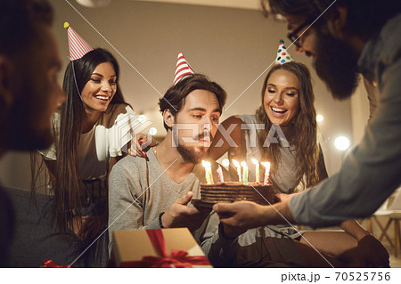 Young man blowing candles on his birthday cake surrounded by happy smiling friends Young man blowing candles on his birthday cake surrounded by happy smiling friends 70525756