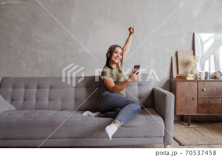 Young brunette woman enjoying the music in wireless headphones connected to smartphone, lying on couch at home alone 70537458