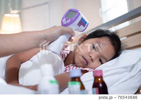 Sick asian child girl is sleeping on bed and her mother is checking up and measuring her body temperature with contactless thermometer in the bedroom at home. 70543686