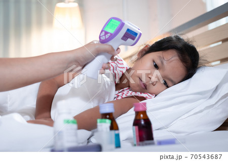 Sick asian child girl is sleeping on bed and her mother is checking up and measuring her body temperature with contactless thermometer in the bedroom at home. 70543687