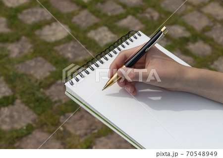 Boy's hand with a yellow-black pen over an open notepad on the background of paving stones and green grass in the park. Clean white sheet. Going to write or draw a picture. The concept of life plannin Boy's hand with a yellow-black pen over an open notepad on the background of paving stones and green grass in the park. Clean white sheet. Going to write or draw a picture. The concept of life plannin 70548949