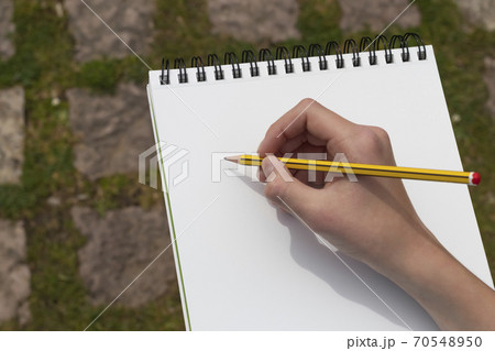 The boy's hand with a yellow-black pencil over an open notepad on the background of paving stones in the park. Clean white sheet. Going to write or draw a picture. The concept of life planning. The boy's hand with a yellow-black pencil over an open notepad on the background of paving stones in the park. Clean white sheet. Going to write or draw a picture. The concept of life planning. 70548950