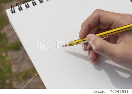 The boy's hand with a yellow-black pencil over an open notepad on the background of paving stones in the park. Clean white sheet. Going to write or draw a picture. The concept of life planning. The boy's hand with a yellow-black pencil over an open notepad on the background of paving stones in the park. Clean white sheet. Going to write or draw a picture. The concept of life planning. 70548951