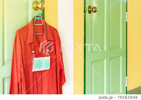 New surgical mask to prevent the spread of Corona Virus(COVID-19) hanging with red shirt on a hanger on a door knob in front of bedroom. New normal lifestyle before go to work concept. New surgical mask to prevent the spread of Corona Virus(COVID-19) hanging with red shirt on a hanger on a door knob in front of bedroom. New normal lifestyle before go to work concept. 70562648