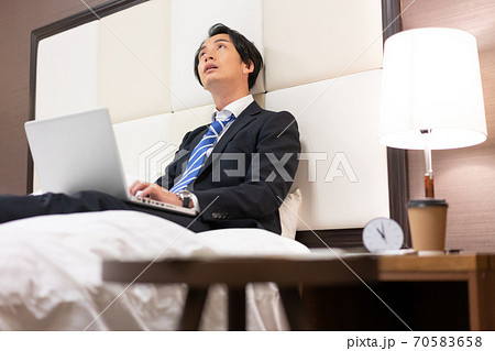 出張先のホテルの部屋のベッドの上でメールチェックをする若いビジネスマン 70583658