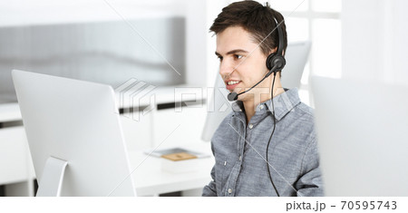 Casual dressed young man using headset and computer while talking with customers online. Call center, business concept 70595743