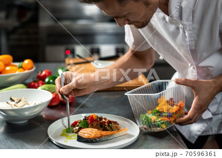 Young male chef decorating roasted vegetables on piece of fried salmon on plate 70596361