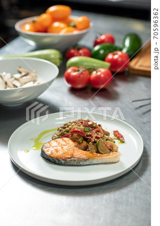 Plate with roasted vegetables on piece of fried salmon standing on kitchen table 70596362
