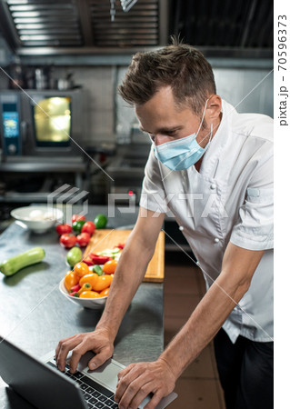 Professional young chef in uniform and protective mask using laptop at work Professional young chef in uniform and protective mask using laptop at work 70596373