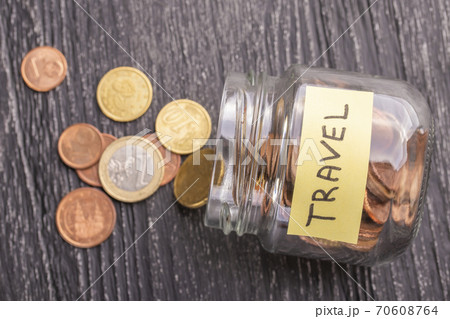 Open tilted glass jar with scattered euro coins with the inscription TRAVEL on a black wooden table. The concept of savings for trips to countries and cities. 70608764
