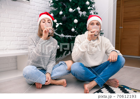 Young happy couple in Christmas hats near a Christmas tree kissing, holding glasses of wine. New Year celebration 70612193