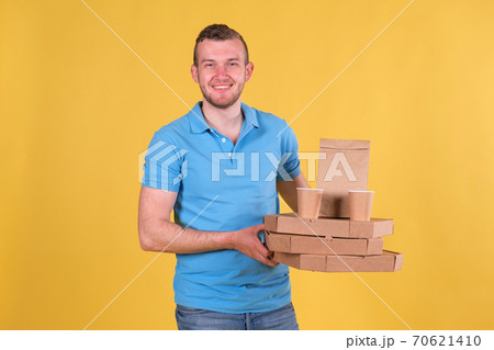 Unshaven young caucasian guy smiling and holding boxes of ready-made food, pizza, coffee, dressed in blue t-shirt and denim pants on yellow background. Concept food grocery delivery for home Unshaven young caucasian guy smiling and holding boxes of ready-made food, pizza, coffee, dressed in blue t-shirt and denim pants on yellow background. Concept food grocery delivery for home 70621410