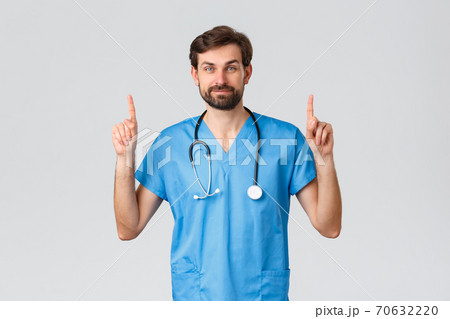 Healthcare workers, pandemic and coronavirus outbreak concept. Handsome bearded doctor in scrubs, stethoscope, pointing fingers up, showing top advertisement, treat patients from covid-19 70632220