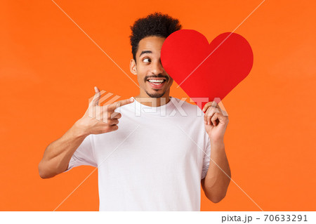 Excited happy, charismatic african-american male in white t-shirt, holding heart valentines card over face, pointing at sign and smiling amused, prepared surprise gift for lover, orange background Excited happy, charismatic african-american male in white t-shirt, holding heart valentines card over face, pointing at sign and smiling amused, prepared surprise gift for lover, orange background 70633291