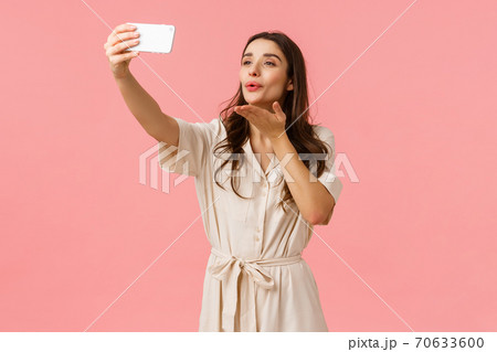 Lovely and tender, alluring brunette female in dress holding smartphone, taking selfie and blowing kiss at mobile camera, stream live video for blog, photographing over pink background Lovely and tender, alluring brunette female in dress holding smartphone, taking selfie and blowing kiss at mobile camera, stream live video for blog, photographing over pink background 70633600