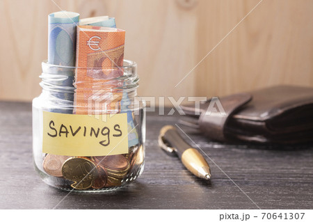 Glass jar with coins and euro notes with the inscription SAVINGS. On the old black wooden table. With a leather wallet and pen. Glass jar with coins and euro notes with the inscription SAVINGS. On the old black wooden table. With a leather wallet and pen. 70641307