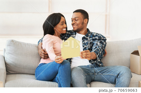 Black millennial woman with her boyfriend hugging and holding paper house in their new apartment, selective focus 70659063