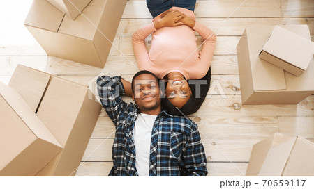 Black young woman with her handsome husband lying among cardboard boxes on floor of their new home, top view Black young woman with her handsome husband lying among cardboard boxes on floor of their new home, top view 70659117