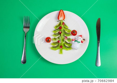 Christmas tree of vegetable and fruit; Kiwi, Strawberry and Pomegranate seed with fork and knife on green background. Merry Xmas and happy New Year party, New You, Healthy food and vegetarian Christmas tree of vegetable and fruit; Kiwi, Strawberry and Pomegranate seed with fork and knife on green background. Merry Xmas and happy New Year party, New You, Healthy food and vegetarian 70662984