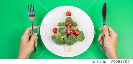 Christmas tree of organic vegetables on white plate; Broccolis and tomatoes with fork and knife on green background. Merry Xmas and happy New Year party, New You, Healthy food and vegetarian concept Christmas tree of organic vegetables on white plate; Broccolis and tomatoes with fork and knife on green background. Merry Xmas and happy New Year party, New You, Healthy food and vegetarian concept 70663209