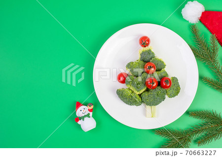 Christmas tree of organic vegetables on white plate; Broccolis and tomatoes with fork and knife on green background. Merry Xmas and happy New Year party, New You, Healthy food and vegetarian concept Christmas tree of organic vegetables on white plate; Broccolis and tomatoes with fork and knife on green background. Merry Xmas and happy New Year party, New You, Healthy food and vegetarian concept 70663227