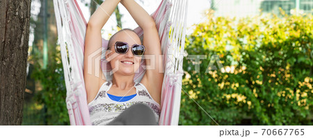 Young adult happy short haired shaved bald attractive woman chilling enjoy relaxing sitting in fabric chair hammock at yard in garden near house. Peaceful and idyllic outdoor rest lifestyle Young adult happy short haired shaved bald attractive woman chilling enjoy relaxing sitting in fabric chair hammock at yard in garden near house. Peaceful and idyllic outdoor rest lifestyle 70667765