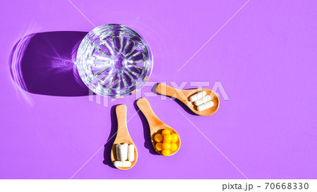 Glass of water and pills on wooden spoon on purple background with hard light and shadows. Creative. Vitamins and prebiotics, probiotics. Dose Glass of water and pills on wooden spoon on purple background with hard light and shadows. Creative. Vitamins and prebiotics, probiotics. Dose 70668330