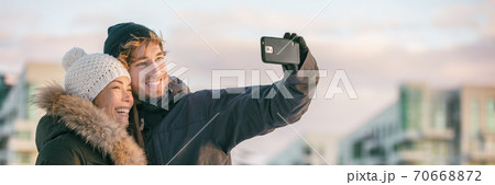 Winter selfie couple tourists on city travel vacation taking photo with mobile phone in street panoramic banner. Happy Asian woman, Caucasian man on holiday 70668872
