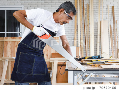 Senior carpenter sawing wood in carpentry workshop. Concept senior people doing hobby after retirement. Senior carpenter sawing wood in carpentry workshop. Concept senior people doing hobby after retirement. 70670466