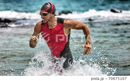 Triathlon swimming man running out of water during ironman race. Male triathlete finishing swim time competition. Fit athlete swimmer sprinting determined out of water in professional tri suit Triathlon swimming man running out of water during ironman race. Male triathlete finishing swim time competition. Fit athlete swimmer sprinting determined out of water in professional tri suit 70675107