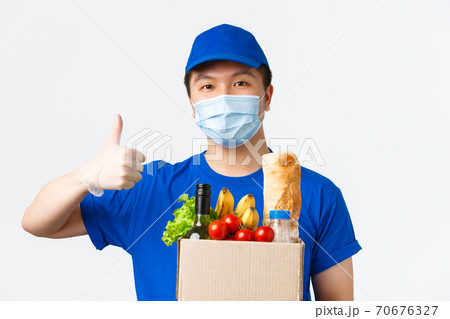 Online shopping, food delivery and covid-19 pandemic concept. Smiling confident asian male courier in medical mask and gloves, blue uniform, carry order, show thumb-up, handing box with groceries Online shopping, food delivery and covid-19 pandemic concept. Smiling confident asian male courier in medical mask and gloves, blue uniform, carry order, show thumb-up, handing box with groceries 70676327