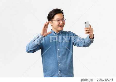 Smiling young asian guy, college student in glasses and braces calling family, using video calling app, waving at smartphone camera to say hi, greeting friend chatting with followers on social media 70676574