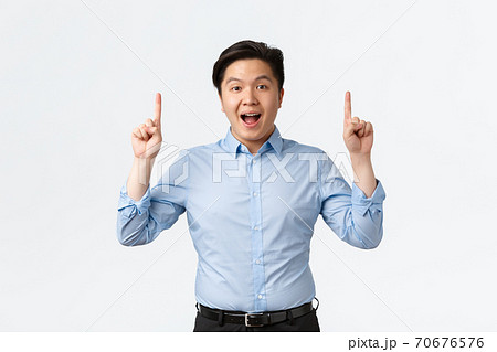 Business, finance and people concept. Excited and amazed asian businessman in blue shirt making announcement, pointing fingers up and look camera, telling big news, white background 70676576