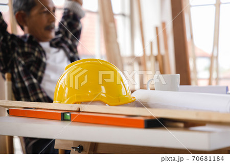 Yellow safety helmet on the table in carpentry workshop with senior carpenter as background. Yellow safety helmet on the table in carpentry workshop with senior carpenter as background. 70681184