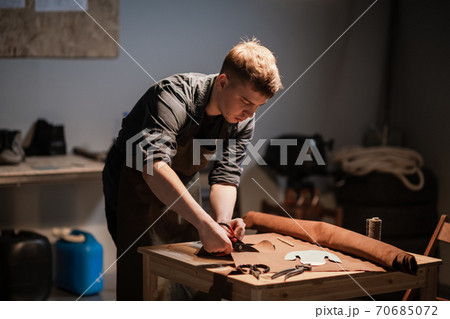 a young shoemaker in a Shoe shop makes shoes from leather with his own hands. 70685072