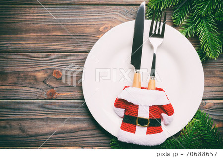 Holiday composition of plate and flatware decorated with Santa clothes on wooden background. Top view of Christmas decorations with empty space for your design. Festive time concept Holiday composition of plate and flatware decorated with Santa clothes on wooden background. Top view of Christmas decorations with empty space for your design. Festive time concept 70688657