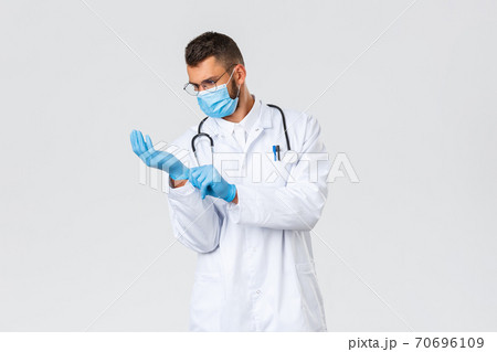 Covid-19, healthcare workers, pandemic, preventing virus concept. Young doctor in white scrubs and medical mask put on rubber gloves to observe patient in clinic, working during coronavirus outbreak 70696109