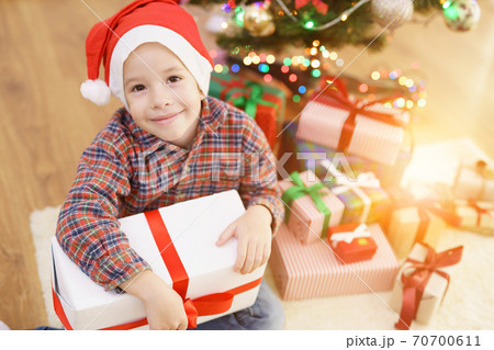 The happy boy sit with a gift box near the christmas tree on a sunny background The happy boy sit with a gift box near the christmas tree on a sunny background 70700611